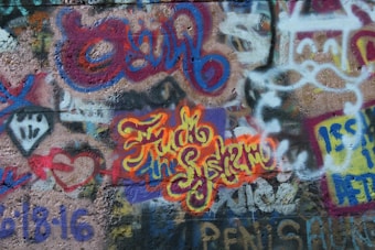 A vibrant and colorful graffiti mural covers a concrete wall. The artwork features overlapping layers of spray-painted text and abstract designs. Bold lettering in yellow and orange stands out with words interwoven in a cursive style. The background includes various other colors like blues, reds, and purples with a mix of geometric and freeform shapes.