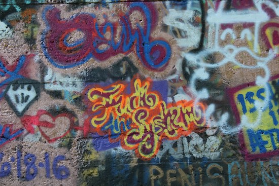 A vibrant and colorful graffiti mural covers a concrete wall. The artwork features overlapping layers of spray-painted text and abstract designs. Bold lettering in yellow and orange stands out with words interwoven in a cursive style. The background includes various other colors like blues, reds, and purples with a mix of geometric and freeform shapes.