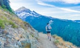 A person is running on a narrow trail along a mountain ridge with a large, snow-capped peak in the background. The landscape features lush green forests and rocky, grassy terrain, under a bright blue sky with wispy clouds.