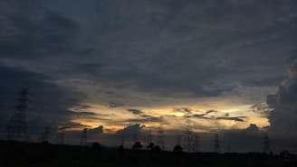 A landscape featuring a dramatic sky filled with dense clouds, with the silhouette of transmission towers stretching across the horizon. The sky is transitioning from day to night, showcasing various shades and patterns as the sun sets.