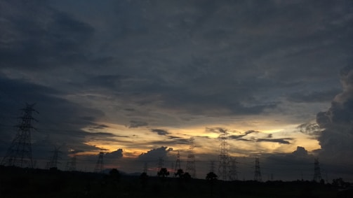 A landscape featuring a dramatic sky filled with dense clouds, with the silhouette of transmission towers stretching across the horizon. The sky is transitioning from day to night, showcasing various shades and patterns as the sun sets.
