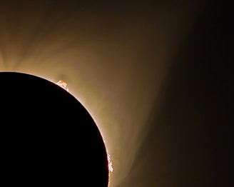A dramatic solar eclipse with the silhouette of the moon partially covering the sun. Glowing solar prominences extend beyond the dark curve, set against a soft, fading gradient of light from golden to brown. The scene captures the celestial phenomenon with a mystical and awe-inspiring ambiance.