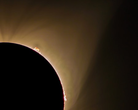 A dramatic solar eclipse with the silhouette of the moon partially covering the sun. Glowing solar prominences extend beyond the dark curve, set against a soft, fading gradient of light from golden to brown. The scene captures the celestial phenomenon with a mystical and awe-inspiring ambiance.