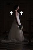A bride in a white wedding gown stands solemnly in a dimly lit room with two illuminated crosses in the background, holding a bouquet of flowers. The floor features a checkered pattern.