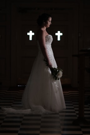 A bride in a white wedding gown stands solemnly in a dimly lit room with two illuminated crosses in the background, holding a bouquet of flowers. The floor features a checkered pattern.