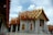 A stunningly ornate temple with intricate golden decorations and red tiled roofs, pillars supporting grand archways, and an expansive marble courtyard with a few statues.