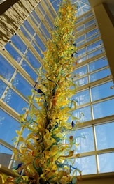 A tall, intricate glass sculpture with vibrant shades of yellow, blue, and green, extending upwards towards a high ceiling with large glass windows. The sculpture's swirling and twisting shapes create a dynamic and artistic impression.