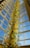 A tall, intricate glass sculpture with vibrant shades of yellow, blue, and green, extending upwards towards a high ceiling with large glass windows. The sculpture's swirling and twisting shapes create a dynamic and artistic impression.
