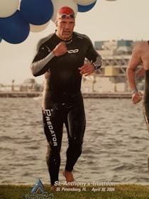 A triathlete in a wetsuit, wearing a swim cap and goggles, is emerging from the water, with droplets scattered around. Blue and white balloons are visible above, and the event identification is printed at the bottom, indicating St. Anthony's Triathlon in St. Petersburg, FL, dated April 30, 2006. In the background, there is a blurred view of a building and more water.