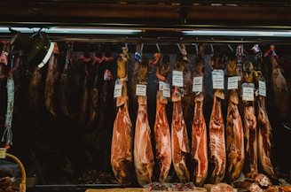 Several cured hams hang from hooks in a shop setting. Labels with prices are attached to each piece, indicating different varieties or brands. The lighting is dim, with a focus on the texture and color of the meat.