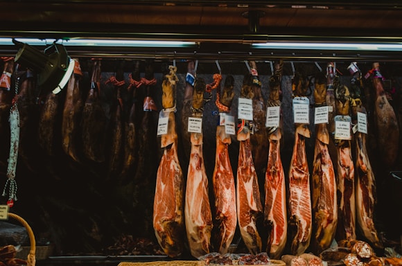 Several cured hams hang from hooks in a shop setting. Labels with prices are attached to each piece, indicating different varieties or brands. The lighting is dim, with a focus on the texture and color of the meat.
