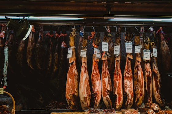 Several cured hams hang from hooks in a shop setting. Labels with prices are attached to each piece, indicating different varieties or brands. The lighting is dim, with a focus on the texture and color of the meat.