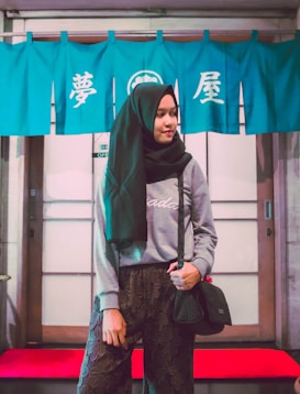 A woman wearing a black hijab and casual attire stands in front of traditional Japanese noren curtains with kanji characters on them. She is carrying a black handbag and appears to be in front of a sliding door with a red doormat, suggesting an entrance to a Japanese establishment.