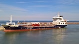A large cargo ship named 'Besiktas Iceland' is docked at a port with calm water. The vessel has a dark hull and white superstructure, with red accents. Behind it, a breakwater protects the harbor, and a distant smaller vessel is visible on the horizon. The sky is clear with light clouds.