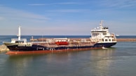 A large cargo ship named 'Besiktas Iceland' is docked at a port with calm water. The vessel has a dark hull and white superstructure, with red accents. Behind it, a breakwater protects the harbor, and a distant smaller vessel is visible on the horizon. The sky is clear with light clouds.