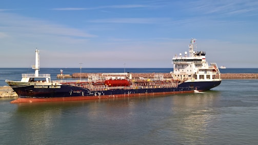A large cargo ship named 'Besiktas Iceland' is docked at a port with calm water. The vessel has a dark hull and white superstructure, with red accents. Behind it, a breakwater protects the harbor, and a distant smaller vessel is visible on the horizon. The sky is clear with light clouds.