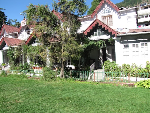 A quaint, rustic house with white walls and red sloped roofs is surrounded by lush greenery. The house features architectural details such as decorative trim under the roof edges and a small, enclosed porch. Potted plants line the border, and a large tree partially obscures the view of the entrance. A well-maintained lawn stretches out in the foreground, and the backdrop includes additional greenery and some neighboring buildings.