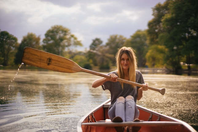 A person sitting in a red canoe or kayak paddling on a calm body of water surrounded by lush green trees. The scene appears serene with sunlight creating a warm, golden glow on the water's surface. The individual is wearing a casual grey shirt and ripped jeans, holding a wooden paddle that is slightly splashing water.