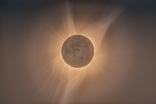 A total solar eclipse with a dark circle blocking the bright sun, creating a halo effect with visible solar corona rays extending outward in a soft, ethereal glow.