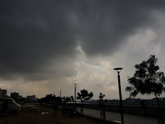 Dark storm clouds dominate a sky over a landscape featuring trees, streetlights, and a pathway. The ground appears wet, reflecting a recent rain or drizzle. In the background, there are outlines of buildings, suggesting an urban setting.