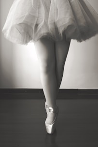 A monochrome image featuring a ballet dancer standing en pointe, showcasing their legs and the lower part of a tutu. The floor appears smooth and dark, contrasting with the lightness of the tutu.
