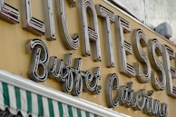 Large, retro-style neon signage displays the words 'Delicatessen Buffet Catering' in bold letters. The sign is mounted on a yellow background. Below the sign, a green and white striped awning is partially visible, suggesting a storefront.