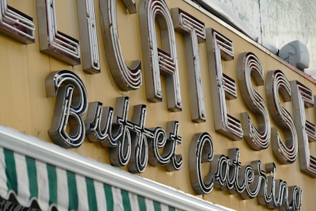 Large, retro-style neon signage displays the words 'Delicatessen Buffet Catering' in bold letters. The sign is mounted on a yellow background. Below the sign, a green and white striped awning is partially visible, suggesting a storefront.