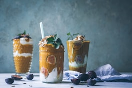 Three layered dessert jars filled with a creamy, orange-colored mixture, topped with fig slices, nuts, and fresh mint. The jars are arranged on a light surface, surrounded by fresh figs and a soft blue fabric, set against a textured gray-blue background.