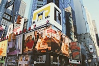 Billboards and advertisements dominate the urban landscape, showcasing products, brands, and entertainers. The scene is bustling with bright signage against the backdrop of towering skyscrapers and city structures.