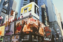 Billboards and advertisements dominate the urban landscape, showcasing products, brands, and entertainers. The scene is bustling with bright signage against the backdrop of towering skyscrapers and city structures.