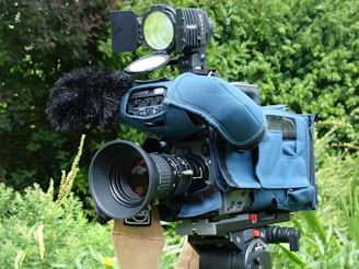 A professional video camera with a large lens and a microphone covered with a windscreen is mounted on a tripod. The camera is encased in a blue protective cover. It is situated outdoors with greenery in the background, including various plants and bushes.