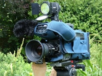 A professional video camera with a large lens and a microphone covered with a windscreen is mounted on a tripod. The camera is encased in a blue protective cover. It is situated outdoors with greenery in the background, including various plants and bushes.