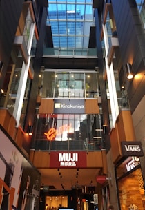 A multi-level shopping space with glass ceilings showcasing several retail stores such as MUJI, Grill'd, and Kinokuniya. The architecture features sleek, modern lines with prominent signage and warm lighting. Reflections on the glass surfaces enhance the sense of depth and space.