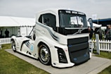 A large, modern, futuristic truck displayed outdoors on a platform. The truck features sleek design lines and is predominantly white with blue and black accents. Its front grille prominently displays the brand name 'VOLVO,' and the words 'THE IRON KNIGHT' are written above the windshield. The scene includes a white picket fence, tents, and several people standing and observing the truck in the background.
