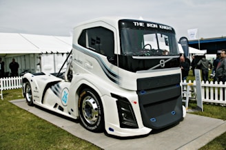 A large, modern, futuristic truck displayed outdoors on a platform. The truck features sleek design lines and is predominantly white with blue and black accents. Its front grille prominently displays the brand name 'VOLVO,' and the words 'THE IRON KNIGHT' are written above the windshield. The scene includes a white picket fence, tents, and several people standing and observing the truck in the background.