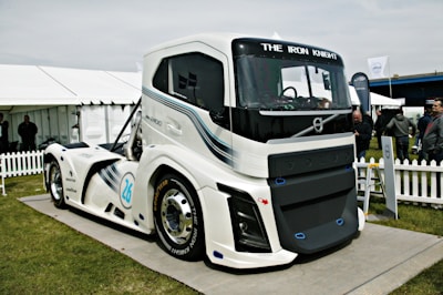A large, modern, futuristic truck displayed outdoors on a platform. The truck features sleek design lines and is predominantly white with blue and black accents. Its front grille prominently displays the brand name 'VOLVO,' and the words 'THE IRON KNIGHT' are written above the windshield. The scene includes a white picket fence, tents, and several people standing and observing the truck in the background.