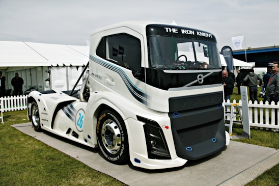 A large, modern, futuristic truck displayed outdoors on a platform. The truck features sleek design lines and is predominantly white with blue and black accents. Its front grille prominently displays the brand name 'VOLVO,' and the words 'THE IRON KNIGHT' are written above the windshield. The scene includes a white picket fence, tents, and several people standing and observing the truck in the background.