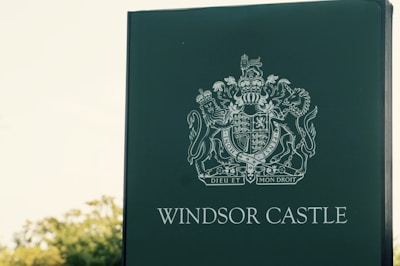 A green sign displaying the Windsor Castle crest with the text 'Windsor Castle' below it. The crest includes intricate designs with symbols such as lions and a crown, along with the motto 'Dieu et mon droit'. The background features some blurred greenery.