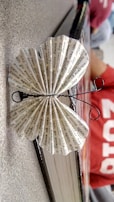 A paper butterfly made from folded printed text is perched on the edge of a book. The text on the paper features repetitive phrases. The butterfly's antennae are crafted from black wire. It is positioned on a textured surface with a person in a red shirt blurred in the background.