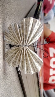 A paper butterfly made from folded printed text is perched on the edge of a book. The text on the paper features repetitive phrases. The butterfly's antennae are crafted from black wire. It is positioned on a textured surface with a person in a red shirt blurred in the background.