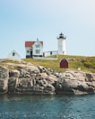 A lighthouse stands on a rocky cliff by the ocean, accompanied by a white house with a red roof and a smaller red shed. The setting is tranquil with clear skies and gentle waves in the water.