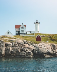 A lighthouse stands on a rocky cliff by the ocean, accompanied by a white house with a red roof and a smaller red shed. The setting is tranquil with clear skies and gentle waves in the water.
