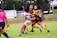 A group of rugby players are actively engaged in a match on a grassy field. One player in a blue and white striped jersey is being tackled by two players in yellow and black striped jerseys. A referee in a pink shirt is nearby, observing the play. Trees and people can be seen in the background, along with a building.