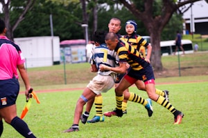 A group of rugby players are actively engaged in a match on a grassy field. One player in a blue and white striped jersey is being tackled by two players in yellow and black striped jerseys. A referee in a pink shirt is nearby, observing the play. Trees and people can be seen in the background, along with a building.
