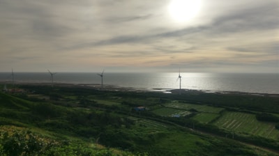 A coastal landscape featuring wind turbines scattered across green fields and hills. The sun is setting or rising over the sea, casting a glow over the water. The sky is partly cloudy with various shades of grey and orange. The overall scene is tranquil and serene, blending natural and man-made elements harmoniously.