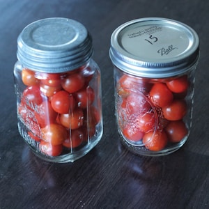 Two glass jars filled with vibrant red cherry tomatoes are placed on a dark wooden surface. Both jars have metallic lids; one lid is plain and the other has a printed label with some text. The jars are simple in design, highlighting the freshness of the tomatoes inside.