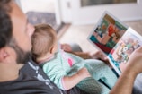 An adult is reading a colorful picture book to a baby who is sitting on their lap. The baby is dressed in pajamas with a cute design. The setting appears to be indoors with soft, natural light illuminating the scene.