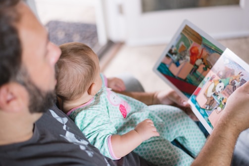 An adult is reading a colorful picture book to a baby who is sitting on their lap. The baby is dressed in pajamas with a cute design. The setting appears to be indoors with soft, natural light illuminating the scene.
