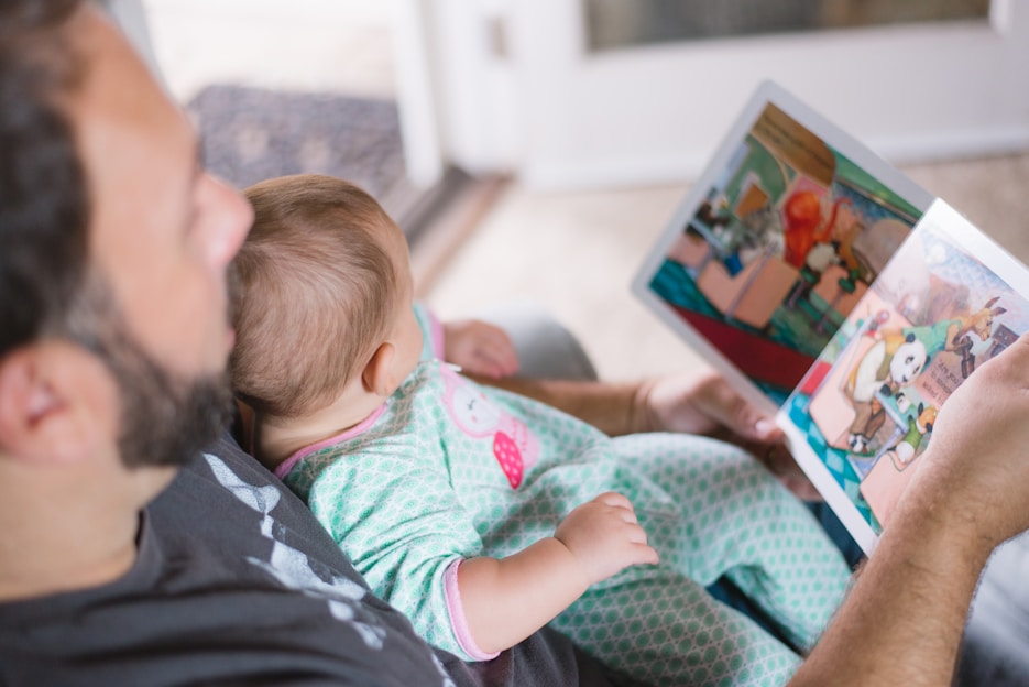 An adult is reading a colorful picture book to a baby who is sitting on their lap. The baby is dressed in pajamas with a cute design. The setting appears to be indoors with soft, natural light illuminating the scene.