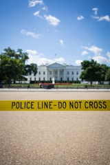 A prominent government building stands in the background, partially obscured by a bright yellow police line with the words 'POLICE LINE - DO NOT CROSS'. The sky is clear with some scattered clouds, and there are trees flanking the building.